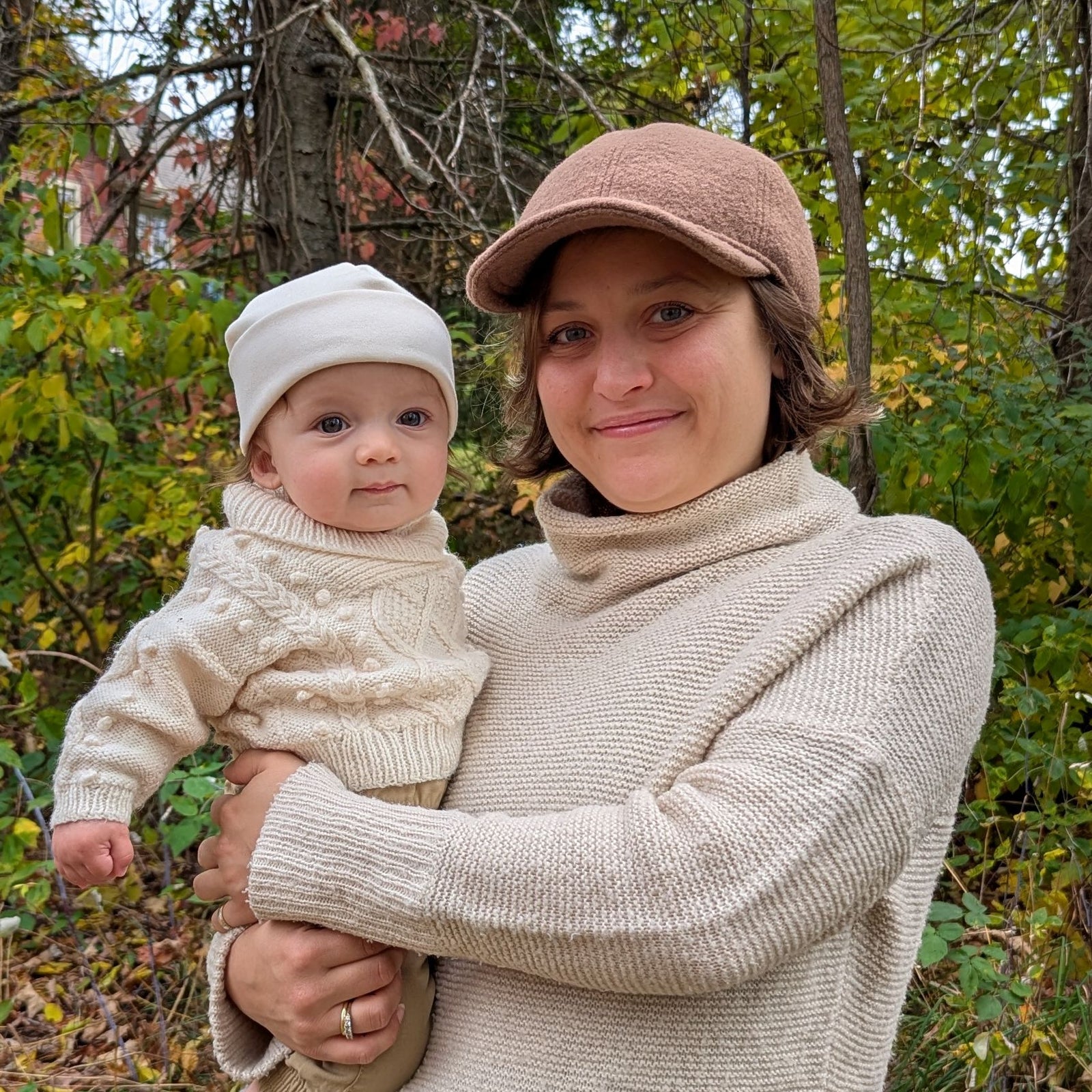 Woman wearing a tan wool ball cap holding infant in irish knit sweater and ivory cotton beanie in autumn woods.