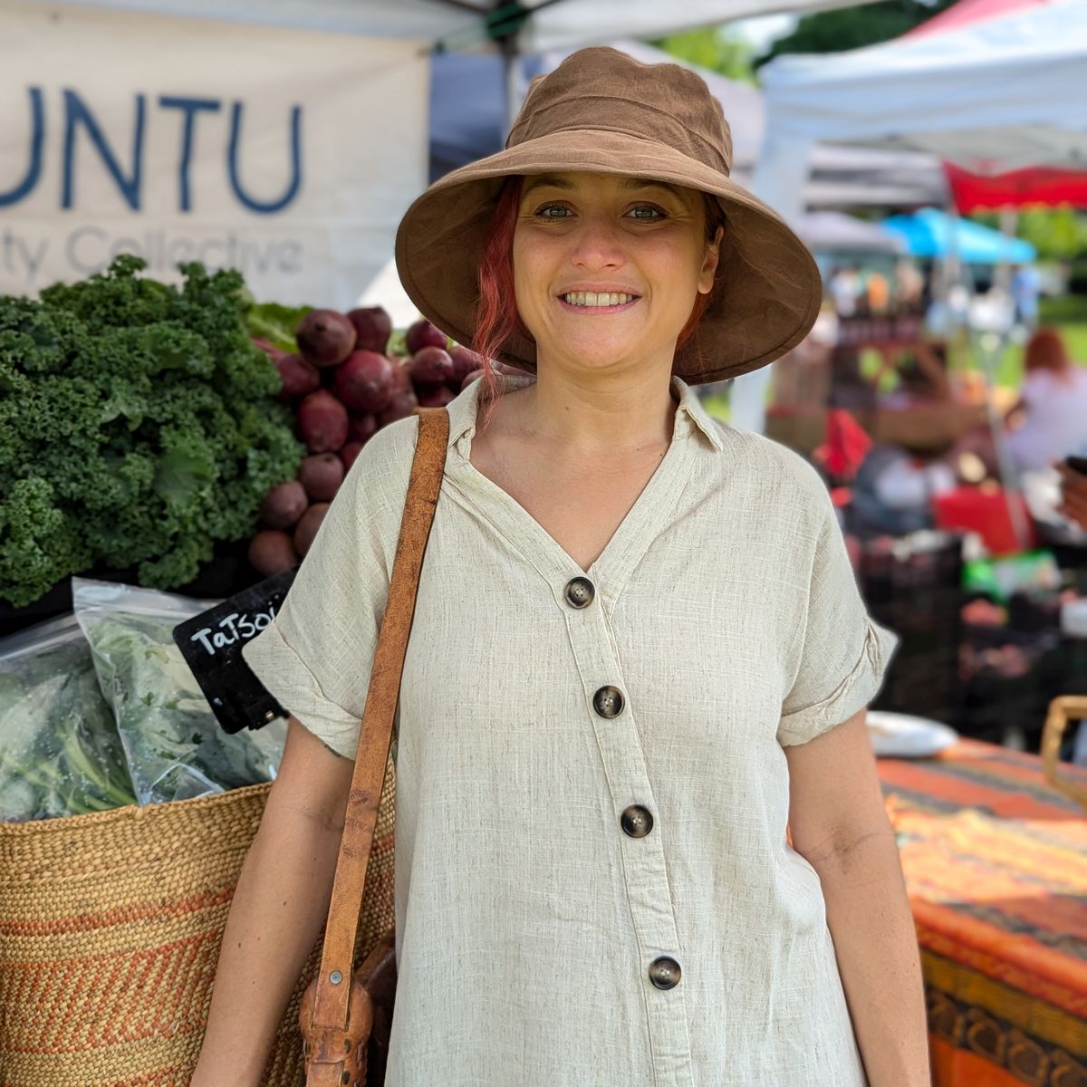 A smiling woman at an outdoor farmers market wearing a light beige linen short-sleeve button-up shirt and a brown wide-brim sun hat, with a woven basket and produce visible in the background.