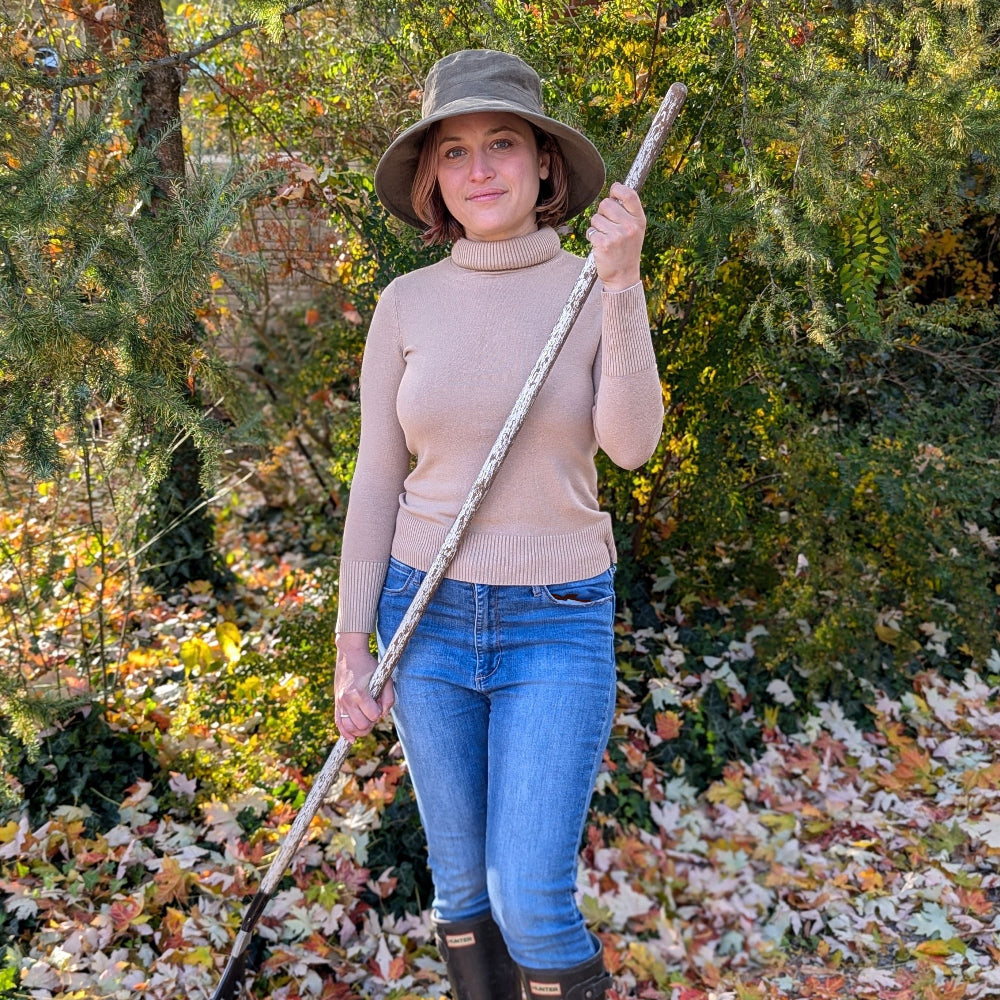Woman raking autumn leaves in jeans, turtle neck and olive green sun hat.