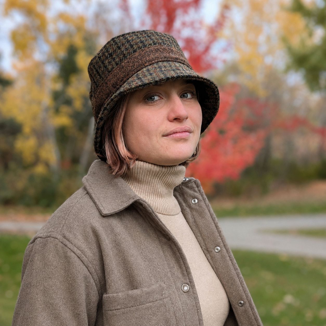 A woman wearing a brown and olive harris tweed wool bucket hat and a taupe button-up shirt over a beige turtleneck, photographed outdoors with a blurred background of autumn trees.