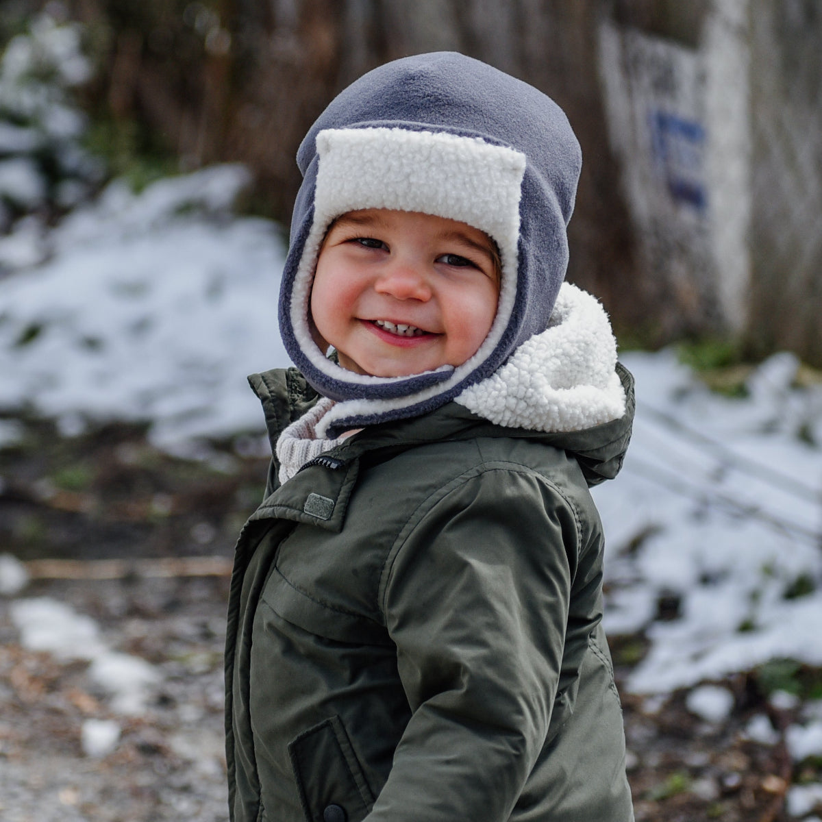 A young child wearing a dark green winter jacket, brown pants, and a gray and white aviator-style winter hat stands outdoors on a snowy path in front of a rustic wooden fence.