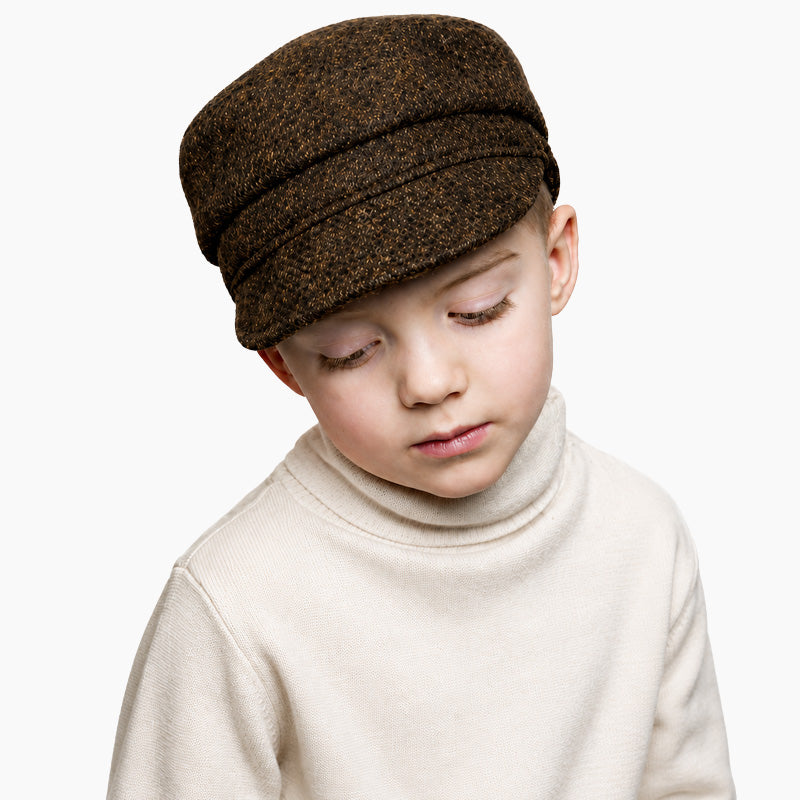 A young child wearing a brown harris tweed cap and a cream-colored turtleneck sweater, photographed against a solid white background.