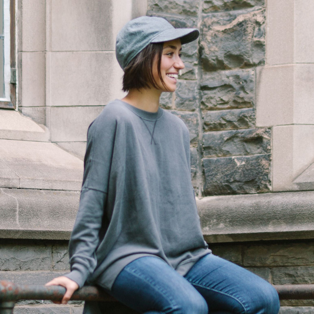 Woman wearing grey linen ball cap sitting on fence rail in front of stone building