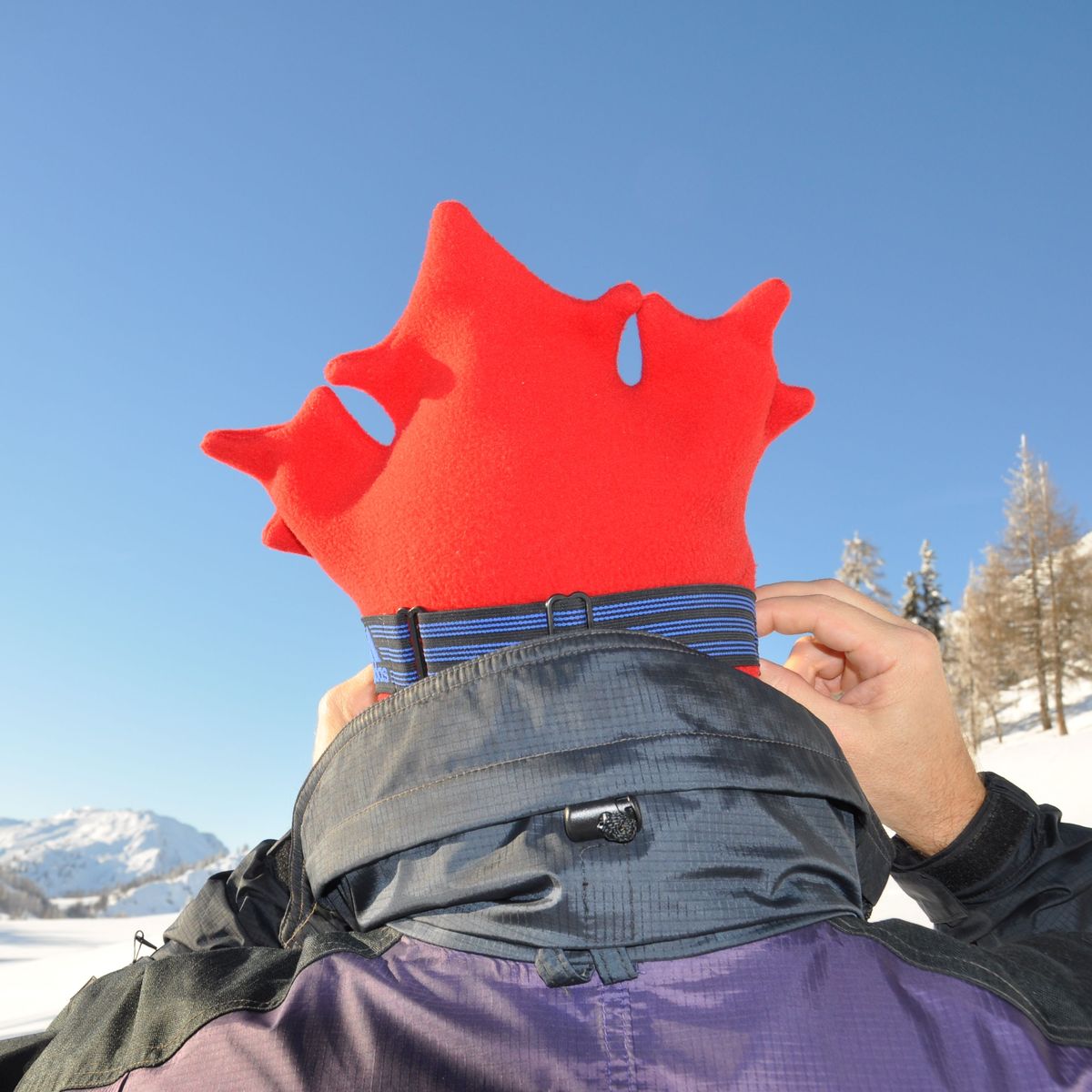 Person on ski hill wearing a red maple leaf shaped toque with google strap and purple jacket