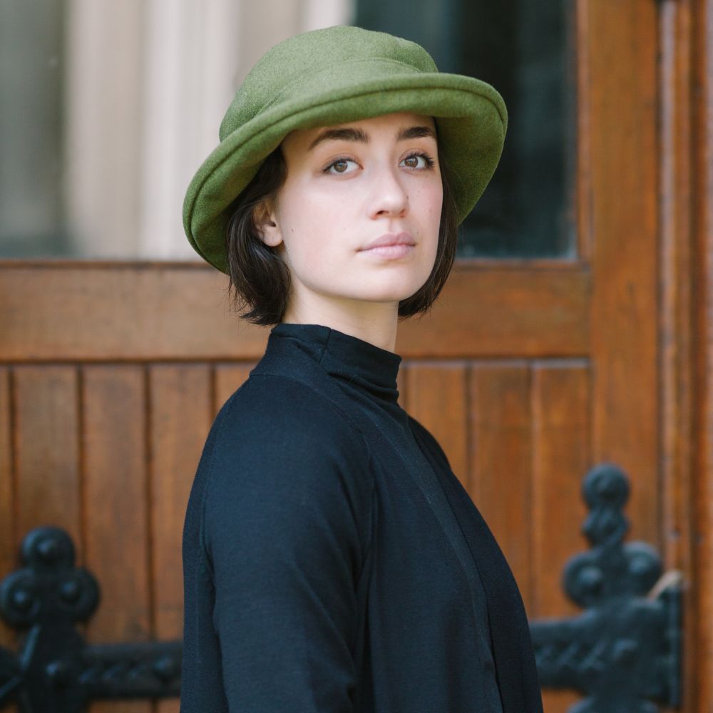Woman in olive green melton wool brimmed bowler hat standing in front of heavy oak door with large black iron hinges