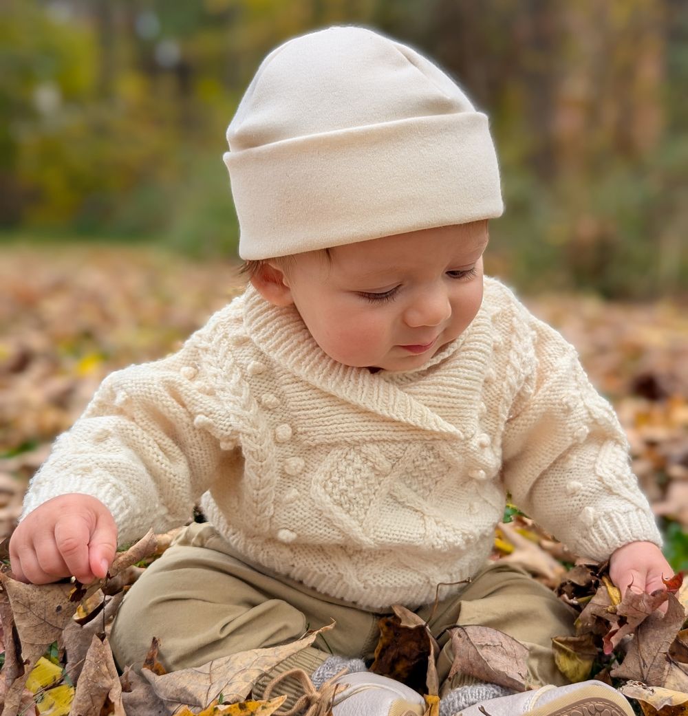 Baby sitting in pile of autumn leaves wearing an irish knit ivory sweater and organic cotton beanie