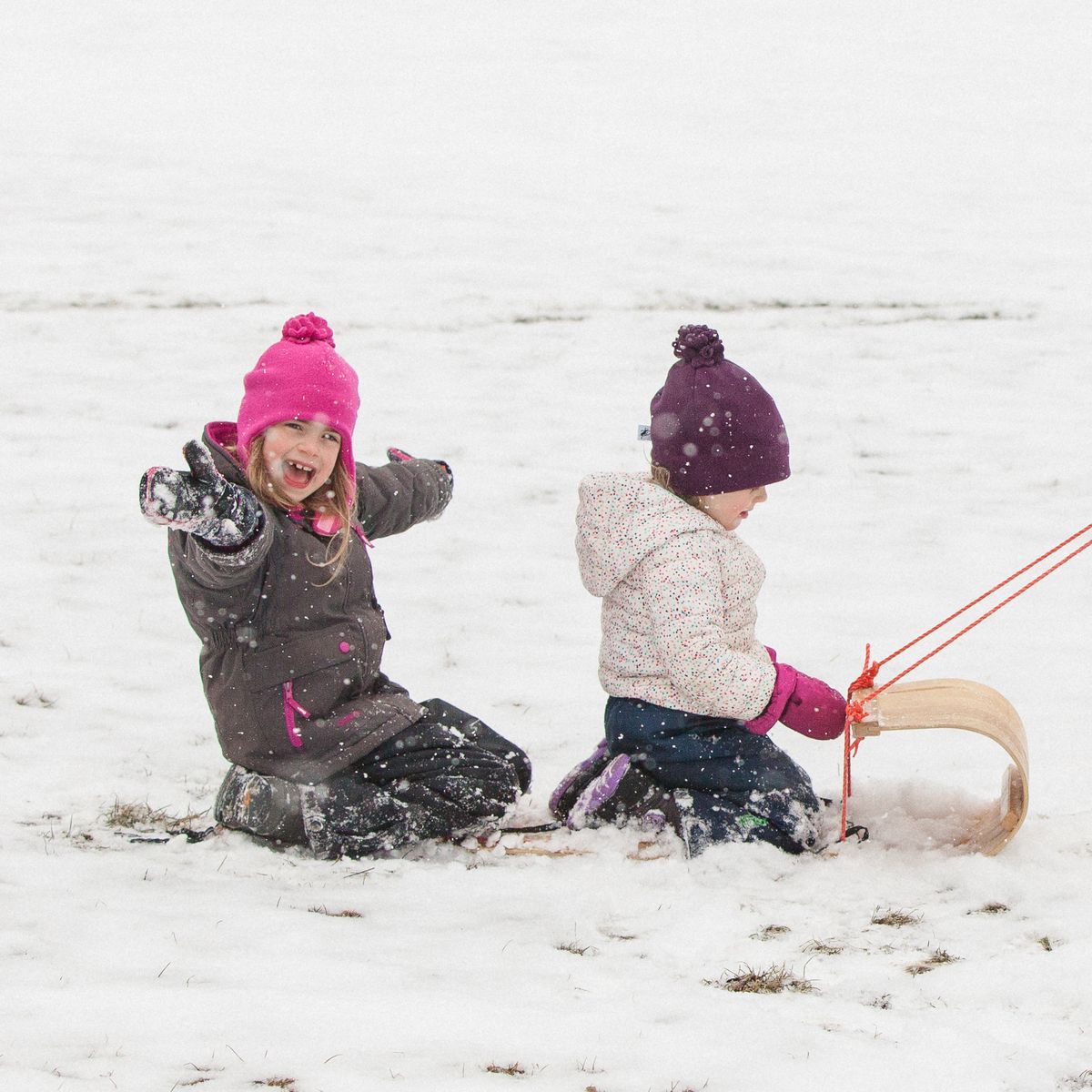 Two kids in Snowsuits and Fleece Hats tobogganing on a snowy day