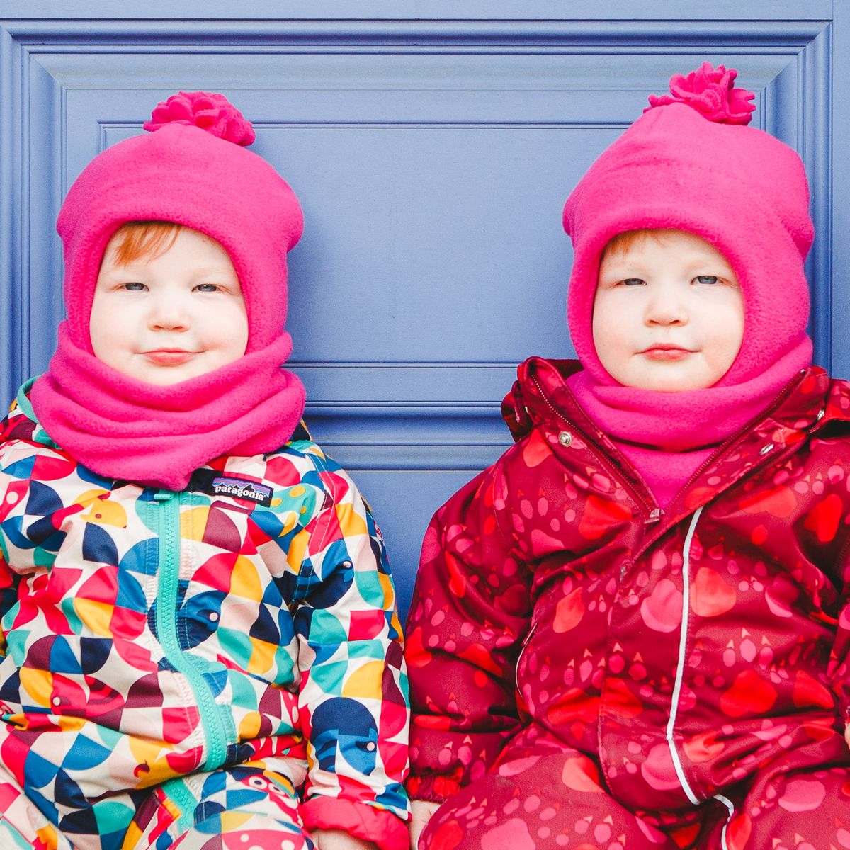 Twins Wearing Snowsuits and Pink Chinwrap Toques with Pompom and Neck Warmers sitting on door step