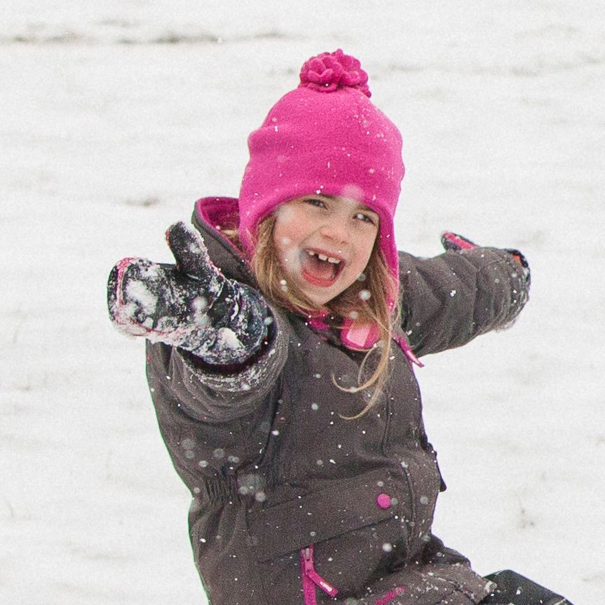 A young girl smiling and throwing snow while wearing a bright pink knit pom-pom beanie, a dark brown snowsuit, and black snow gloves in the snow.