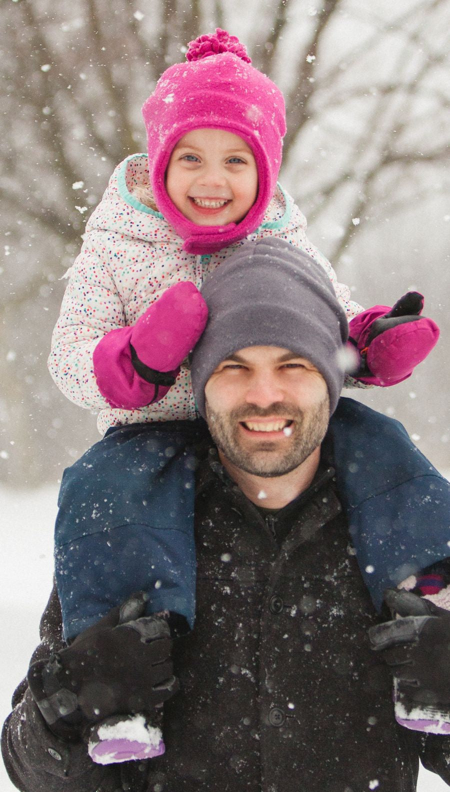 Man with child on shoulders wearing toques walking through snowy park