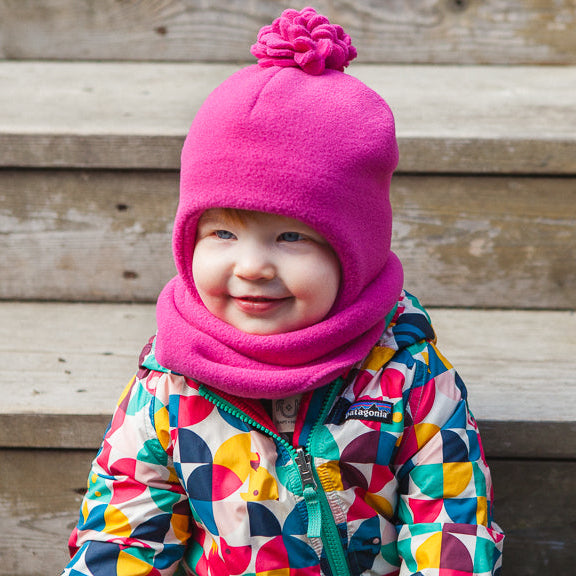 A smiling young child wearing a bright pink fleece bonnet with a pom-pom and matching pink neck warmer, paired with a colorful geometric print winter jacket, posed in front of rustic wooden steps.