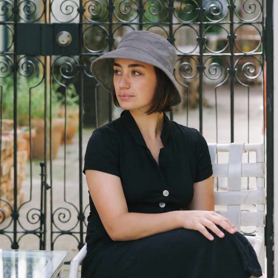Woman wearing grey 3 inch brim Linen bowler hat, sitting on patio with iron gate in background