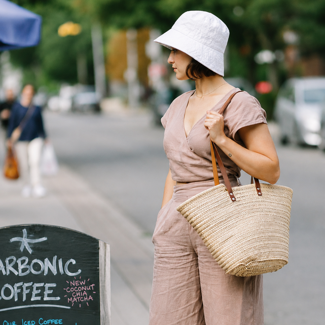 A woman wearing a Puffin Gear white linen bowler hat in front of coffee shop carrying a market bag.