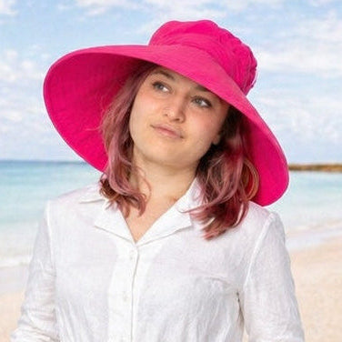 Woman wearing an ultra wide 6 inch brim bright pink hat with white shirt standing on beach.