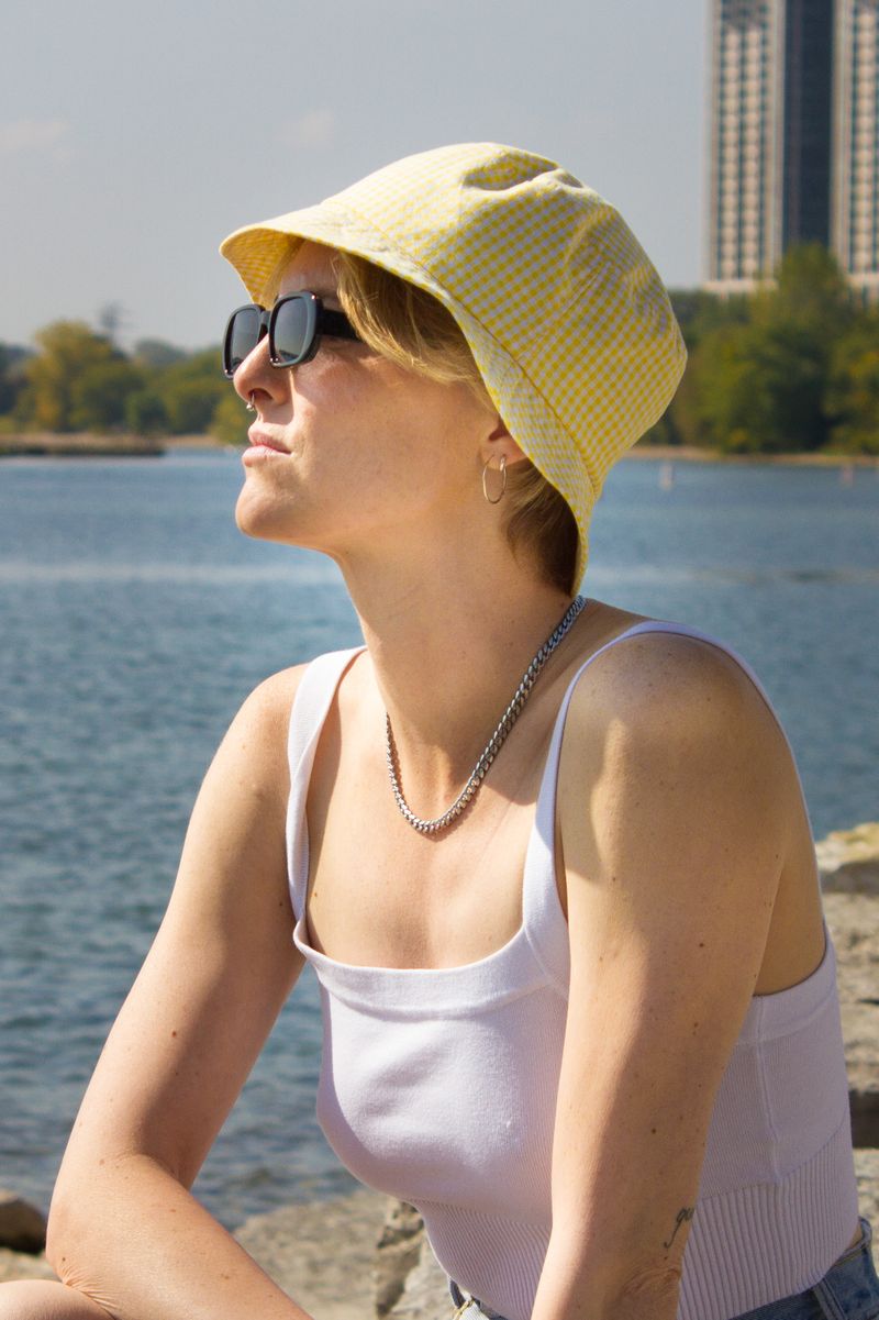 A woman wearing a light yellow gingham sun hat, black sunglasses, and a white ribbed tank top sits outdoors by the water with a city skyline in the background. UPF50 sun protection made in Canada by Puffin Gear