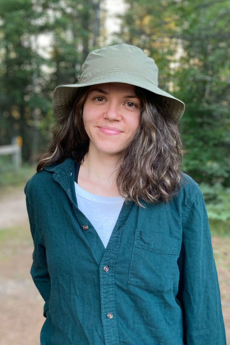 A woman wearing an olive green wide-brim bucket hat and a dark teal button-up shirt, standing outdoors in a wooded setting. UPF50 sun protection made in canada by puffin gear.