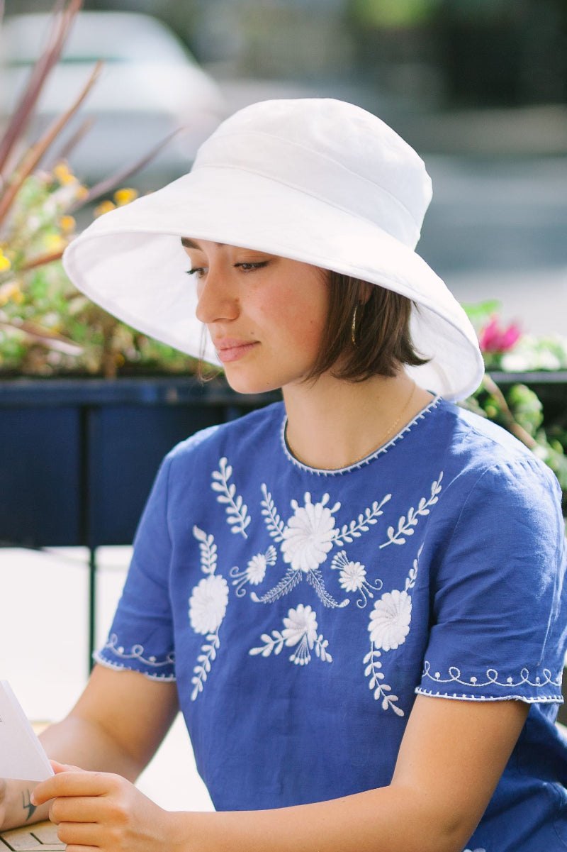 A woman wearing a white linen wide-brim sun hat and a blue short-sleeve top with white floral embroidery, looking down while holding reading a menu on a patio